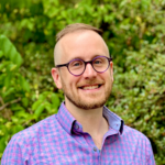 A person with a beard and glasses smiling in front of green foliage, wearing a purple checkered shirt.