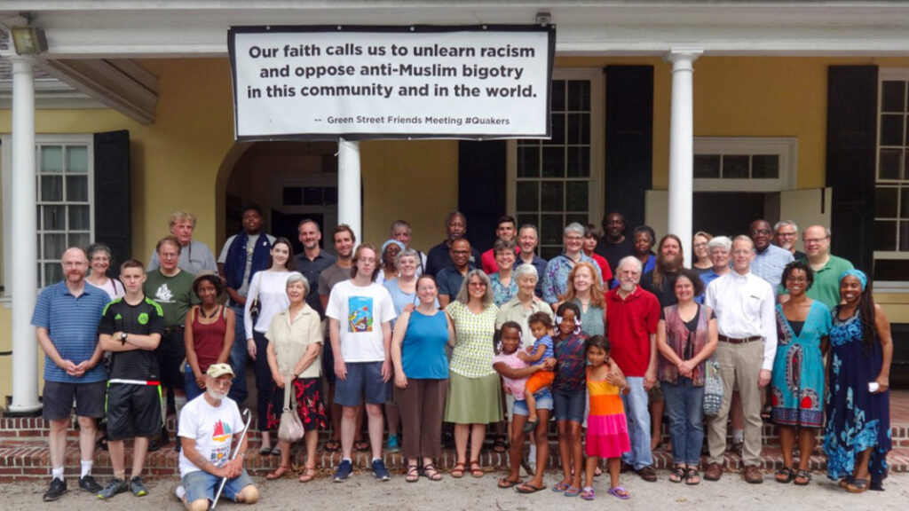 A diverse group poses in front of a building with a sign promoting racial and religious inclusion.