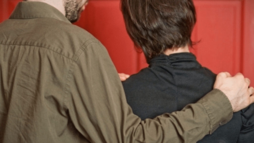 Two people in dark clothing from the back, one with their arm around the other