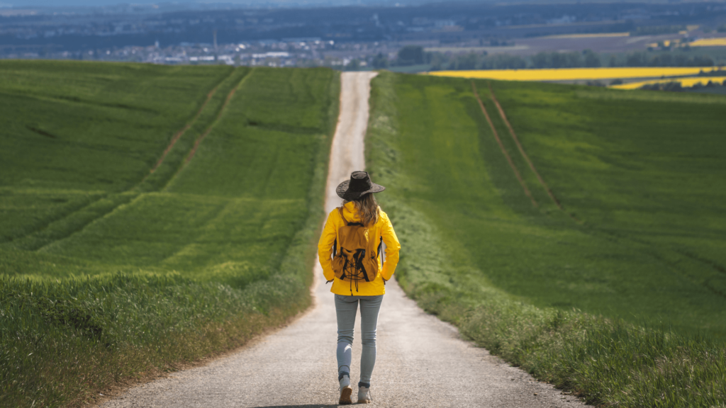 A woman in a yellow jacket and brown hat walking down a road with green fields on either side