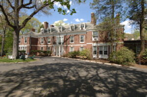 A large, three-story brick building with white trim, surrounded by trees and a paved driveway.