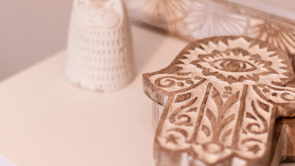 A carved wooden Hamsa hand rests on a table next to a small decorative ceramic figure.