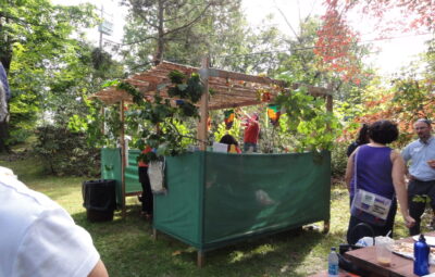 People gathered around a green booth with foliage at an outdoor event on a sunny day.