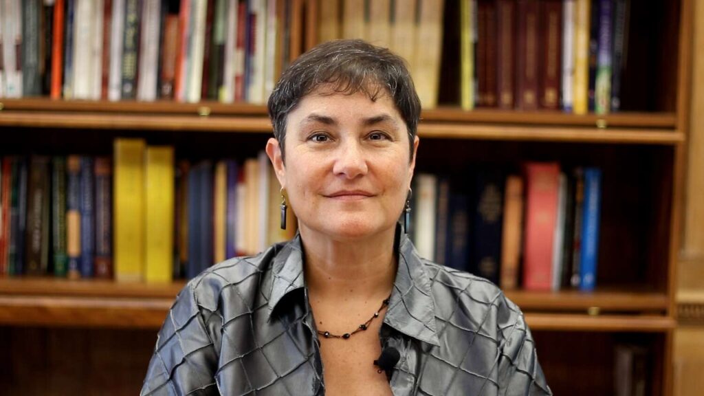 Person with short hair and earrings in front of bookshelves, wearing a gray shirt and necklace.