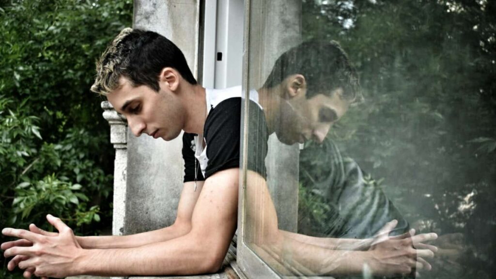 Young man sitting with head down and hands together with reflection in glass window