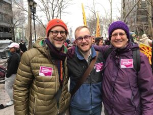 Three people smiling and standing together at an outdoor event, wearing winter clothing and pink stickers.