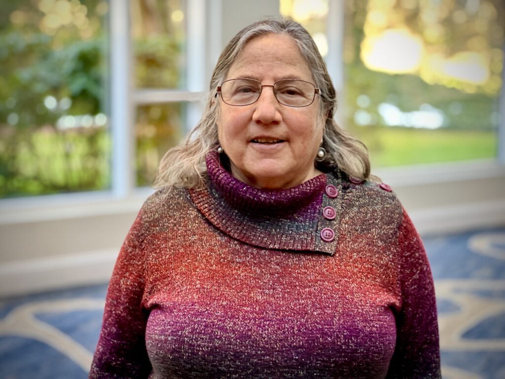 A waist up photo of Jackie Land. She's wearing a red sweater, standing in front of floor-to-ceiling windows.