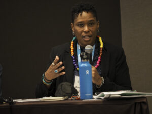 Person speaking at a panel, wearing colorful beads and holding a microphone near a blue water bottle.