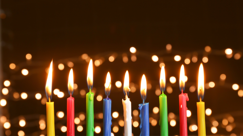 Colorful lit candles in a row with bright, blurred lights in the background.
