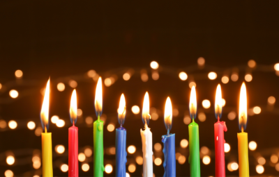 Colorful lit candles in a row with bright, blurred lights in the background.