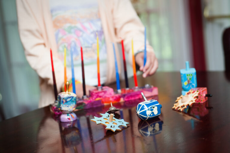 A reddish, pink menorah with multicolored candles rests on a table, with a smattering of dreidels. Someone wearing a pink sweater stands behind, putting the eight candle in.