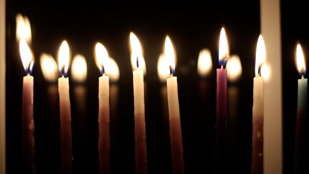 A row of lit candles in various colors against a dark background.