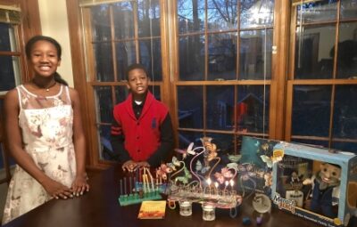 Two children smile near a table with a lit menorah, a magic set, and festive decorations.