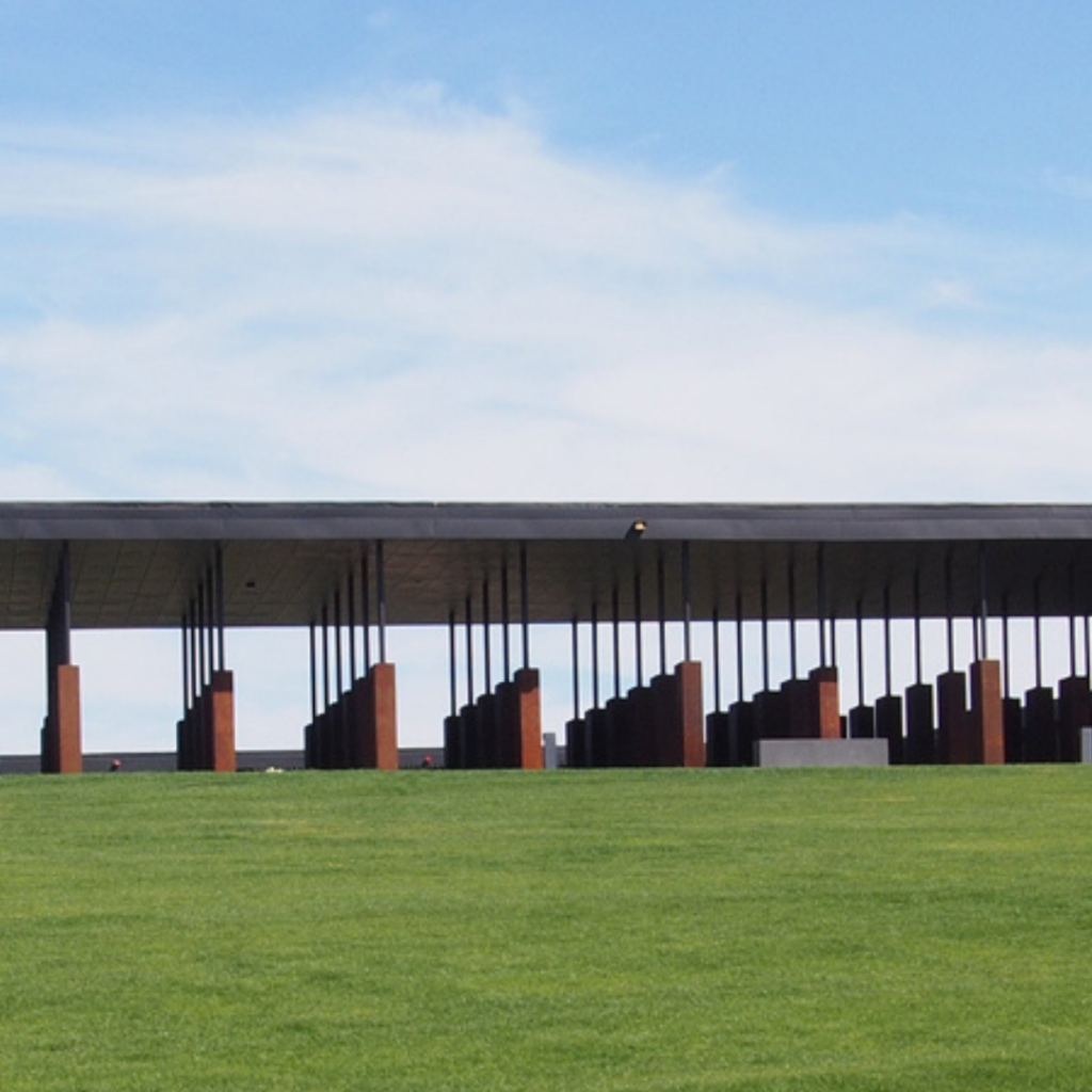 Modern building with a flat roof and tall, vertical supports on a green lawn under a blue sky.