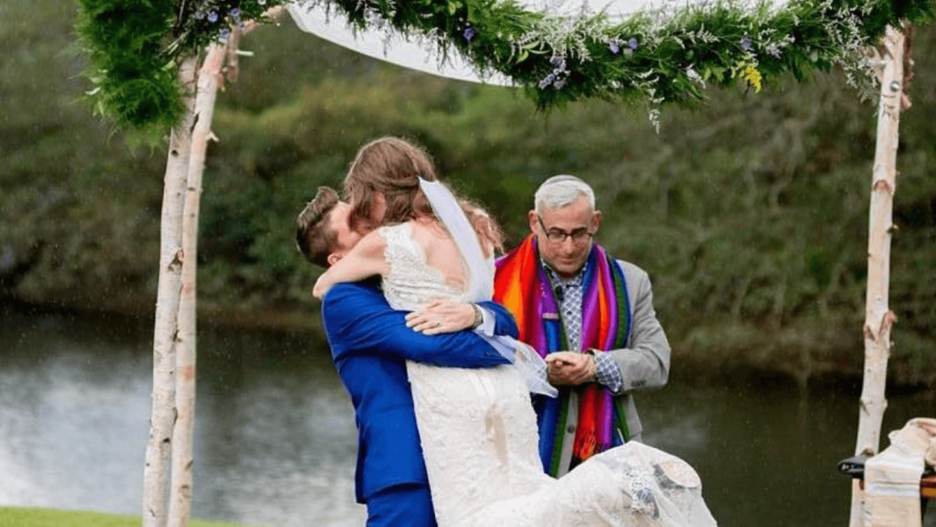 Two brides kissing under a chuppah with a rabbi in the background