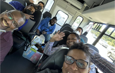 A group of people smiling and waving inside a bus. Some are wearing masks. There are supplies in the aisle.