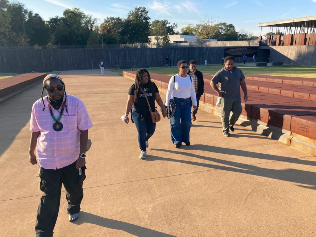 Walking four abreast, not not exactly parallel, participants in the pilgrimage take in the National Memorial for Peace and Justice, beneath by a blue sky.