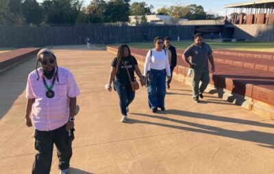 Walking four abreast, not not exactly parallel, participants in the pilgrimage take in the National Memorial for Peace and Justice, beneath by a blue sky.