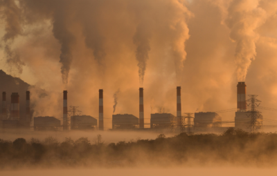 Row of smokestacks releasing smoke into an orange, hazy sky, with power lines and trees in the foreground.
