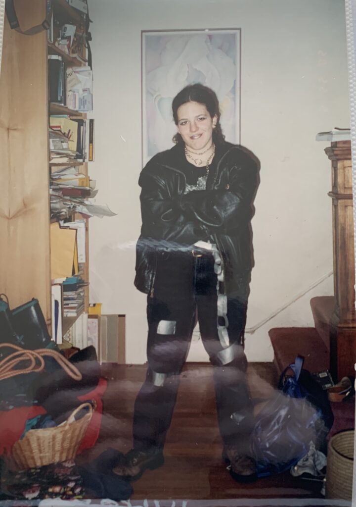 Person in black leather jacket and pants stands in a cluttered room with shelves, books, and a large painting behind.