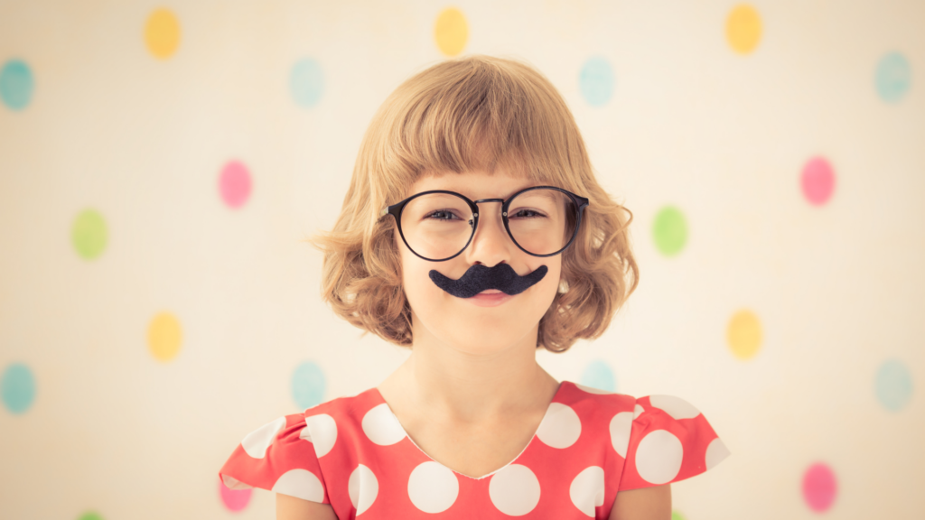 Child in polka dot dress wearing glasses and a fake mustache, with colorful polka dot background.