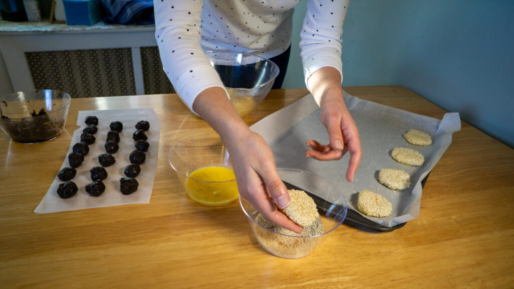 Person coating cookies in crumbs, with chocolate balls and batter on a wooden table.