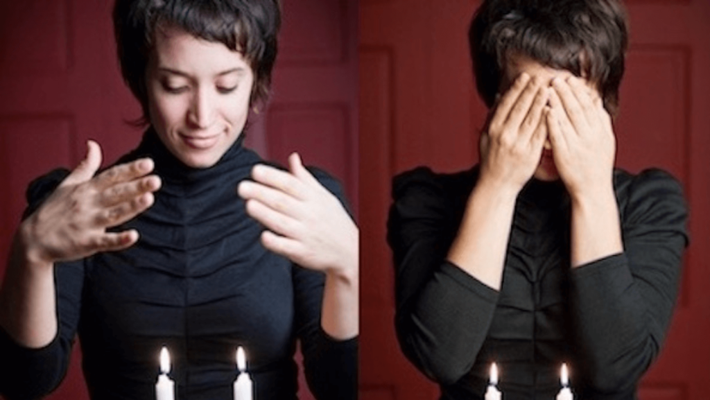 A woman with short dark hair covering her eyes in front of lit Shabbat candles