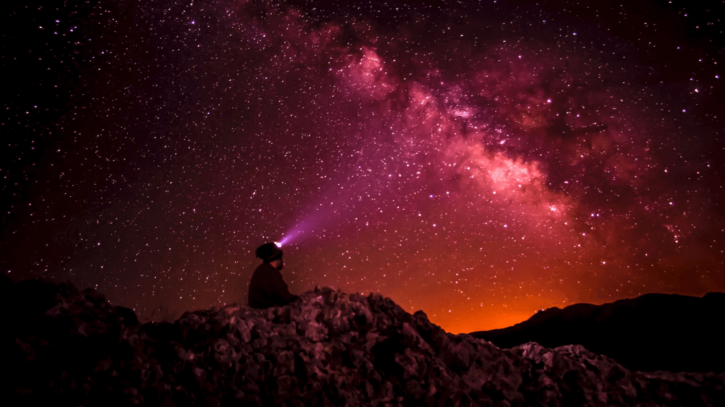 Person in a headwrap sitting on a hill looking up at a pink, purple, and orange night sky
