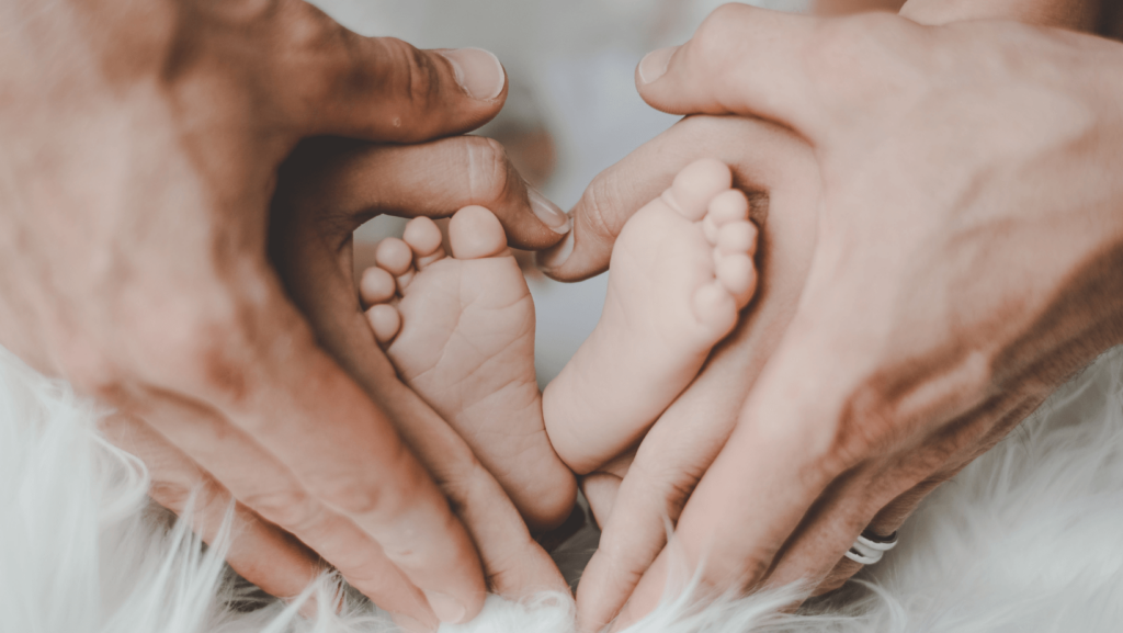 Two adult sets of hands forming a heart shape around a pair of baby feet