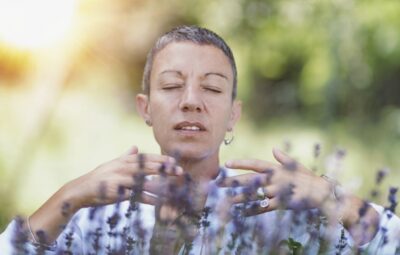 Person with short hair meditates outdoors among lavender plants, eyes closed in a serene natural setting.