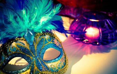 A decorative blue and gold masquerade mask with feathers next to a lit purple candle on a white surface.