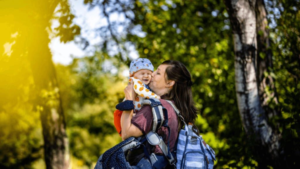 A mother holding and kissing a baby outside near trees