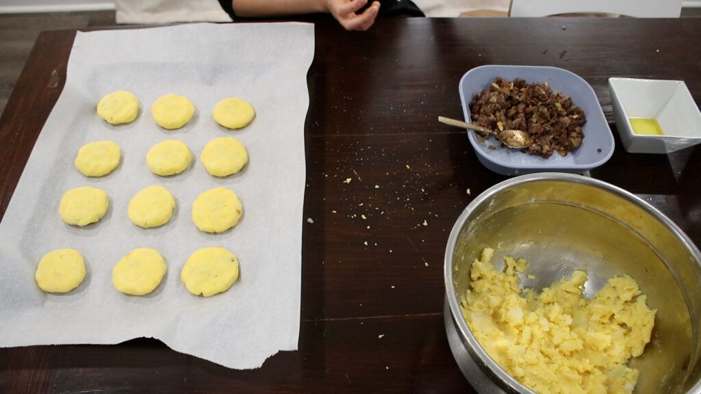 Dough balls on parchment with meat filling and mashed potatoes on a dark table.