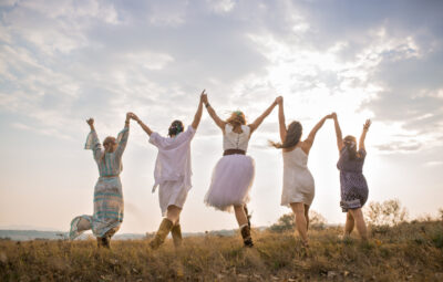 Five people in dresses hold hands and celebrate under a partly cloudy sky in an open field.