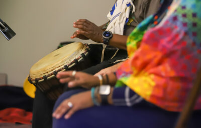 Two people are sitting, playing a hand drum. One wears a colorful outfit, and both have watches and bracelets.