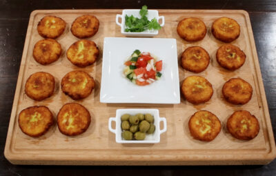 A wooden board with patties, olives, salad, and greens neatly arranged.