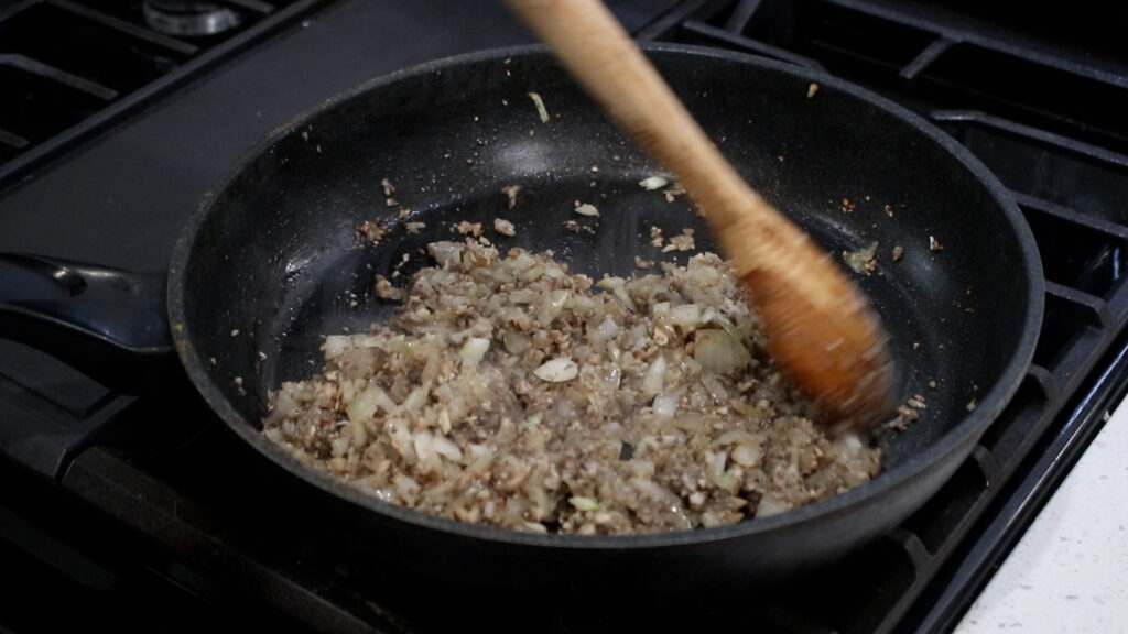 Chopped onions and ground beef cooking in a black pan, stirred with a wooden spoon.
