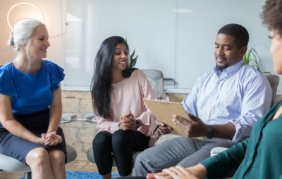 Four people having a discussion in an office setting, seated in a semi-circle with a clipboard.