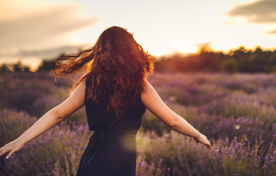 A woman with curly hair in a black dress dances freely in a lavender field at sunset.