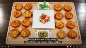 A wooden board with kubbah, a central plate of salad, and a small bowl of olives. Subtitles describe Passover traditions.
