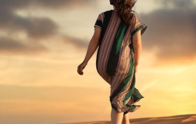 Person in a striped dress walking barefoot on sand dunes at sunset, with clouds in the sky.