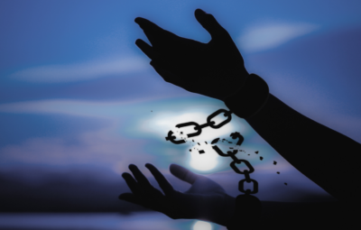 Silhouetted hands breaking free from chains against a blue sky with a glowing light in the background.