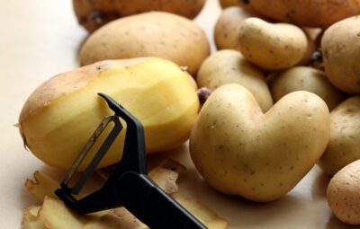 Peeled potato and peeler among several unpeeled potatoes on a wooden surface.