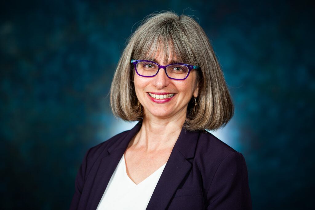 Rabbi Mira Wasserman, stands in front of a blue screen.