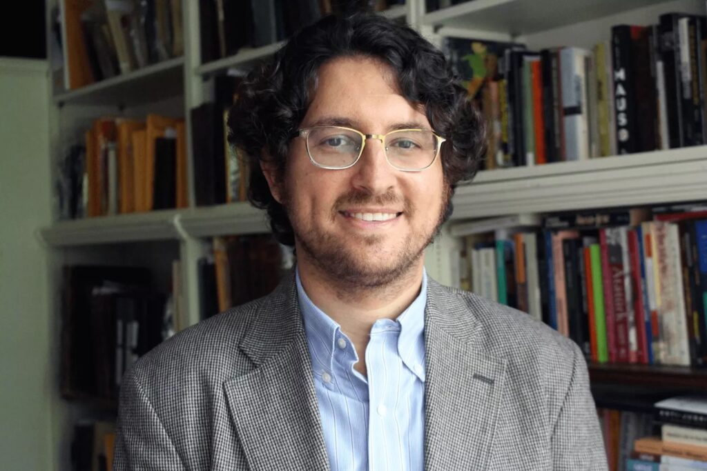 A man with glasses and curly hair smiles, standing in front of a bookcase filled with various books.