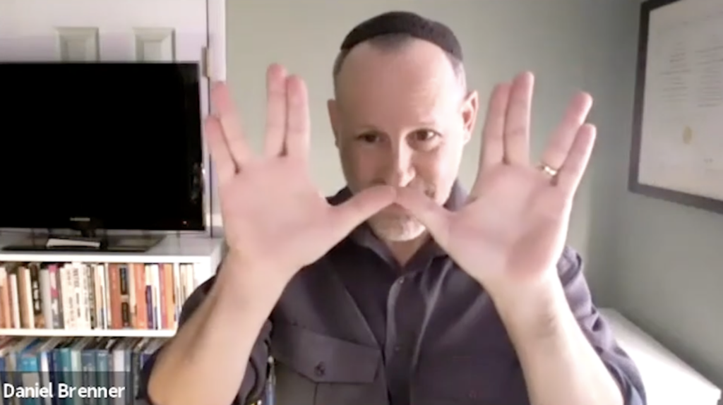 A man in a dark shirt makes a Vulcan salute with both hands in a room with a TV and bookshelves.
