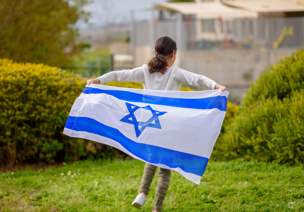 Person holding an Israeli flag, walking in a grassy area with bushes in the background.