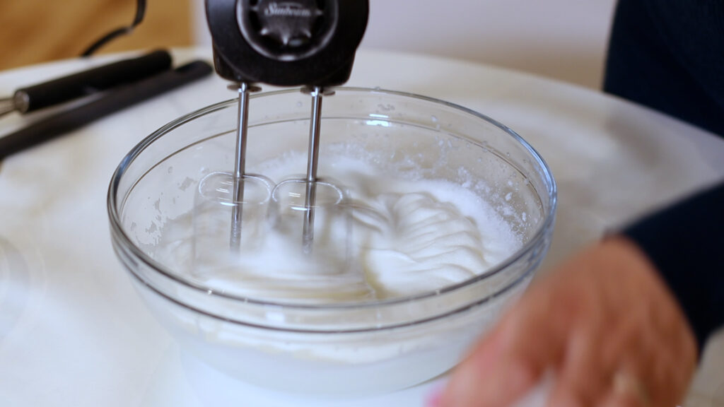 Electric hand mixer whipping egg whites in a glass bowl on a white surface.