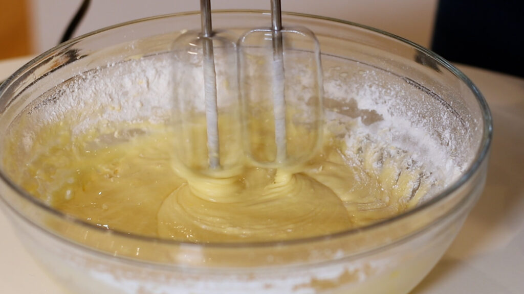 Electric beaters mixing yellow cake batter in a glass bowl.