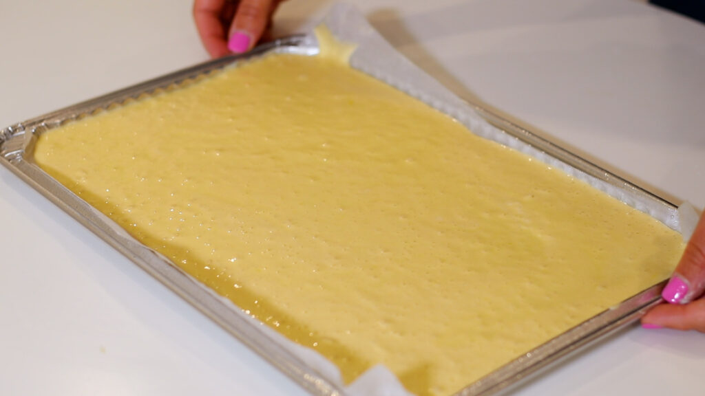 Hands holding a baking tray with a layer of uncooked yellow batter spread evenly on parchment paper.